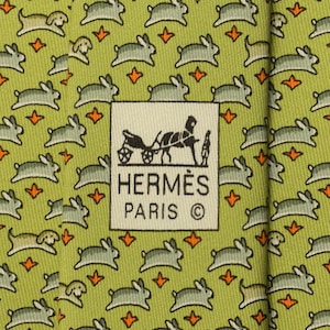 May include: A light green necktie featuring a repeating pattern of small grey rabbits and tiny orange star-like shapes. A central white rectangular label displays a black graphic of a horse-drawn carriage with the text HERMÈS PARIS ©.
