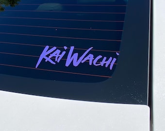 May include: A purple "Kai Wachi" decal is affixed to the rear window of a white vehicle. The decal is in a stylized font and is centered on the glass. The background shows the interior of the car and a glimpse of the outside.