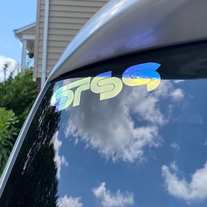 May include: A holographic sticker with the letters "SRSG" in a stylized font is affixed to a car window. The sticker reflects a rainbow of colors. The sky and clouds are reflected in the window.