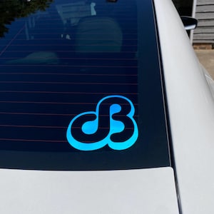 May include: A blue and black decal with a stylized "dB" design is affixed to the rear window of a white car. The decal has a gradient effect, transitioning from a lighter blue to a darker blue.