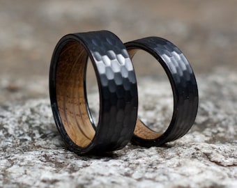 May include: Two black hammered wedding bands with a wood inlay. The bands are on a gray rock surface.