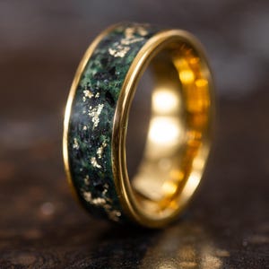 May include: A gold ring with a dark green inlay speckled with gold flakes. The ring has a polished gold band and a smooth, reflective surface. The ring is a piece of jewelry.