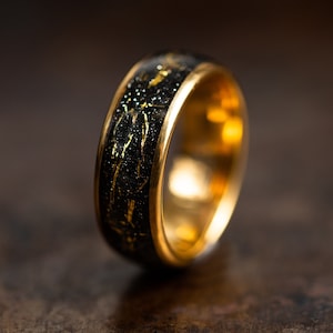 K&ouml;nnte beinhalten: Ein Goldring mit einer schwarzen Einlage mit goldenen Flocken. Der Ring hat eine polierte Goldoberfl&auml;che.