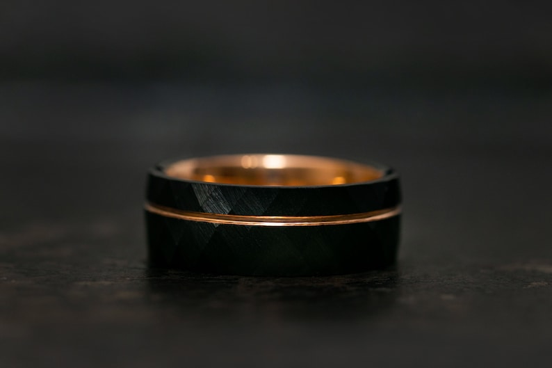 Puede incluir: Un anillo de carburo de tungsteno negro con una banda de oro rosa. El anillo tiene un patr&oacute;n de corte de diamante.