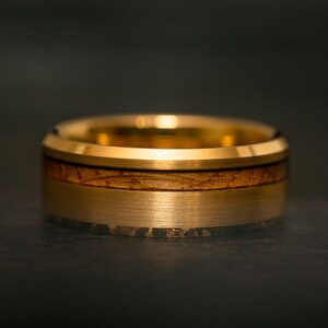 May include: Close-up of a gold-colored ring with a wood inlay. The ring has a brushed metal finish on the outer band and a polished gold edge. The wood inlay is a light brown color.
