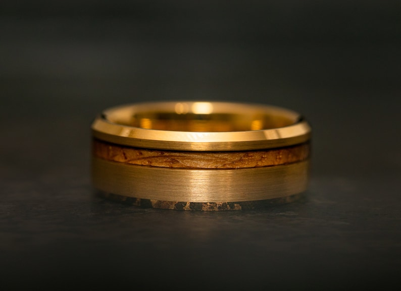 May include: Close-up of a gold-colored ring with a wood inlay. The ring has a brushed metal finish on the outer band and a polished gold edge. The wood inlay is a light brown color.