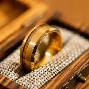May include: A gold-colored ring with a wood inlay, presented in a wooden box. The ring features a brushed metal finish and a band of light brown wood. The box has a rustic aesthetic, with a woven fabric lining.