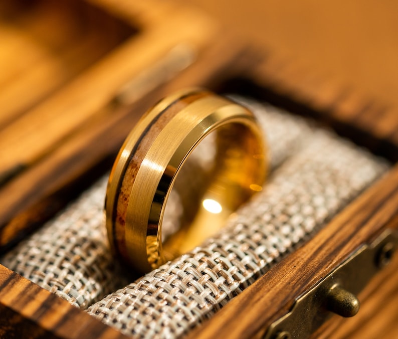 May include: A gold-colored ring with a wood inlay, presented in a wooden box. The ring features a brushed metal finish and a band of light brown wood. The box has a rustic aesthetic, with a woven fabric lining.