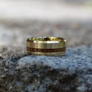 May include: A gold-colored ring with a brushed finish and a band of dark brown wood inlay. The ring is set against a neutral, textured background, highlighting its design and craftsmanship. A unique piece of jewelry.