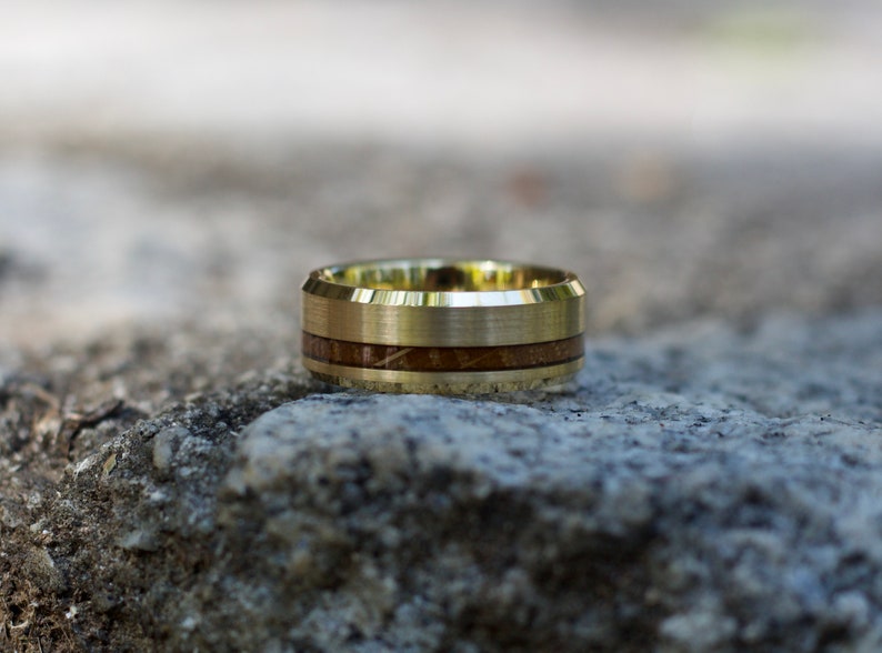 May include: A gold-colored ring with a brushed finish and a band of dark brown wood inlay. The ring is set against a neutral, textured background, highlighting its design and craftsmanship. A unique piece of jewelry.