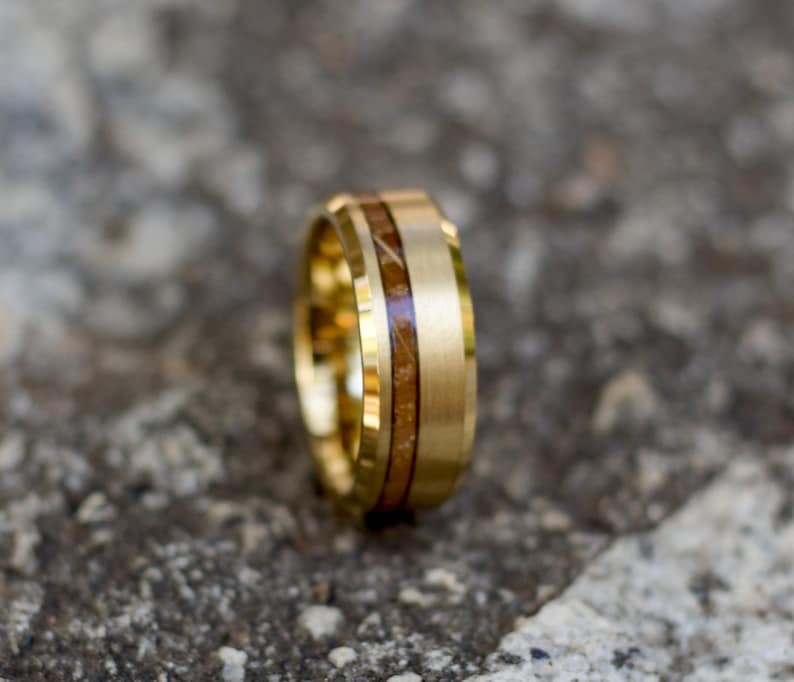 May include: A gold-colored ring with a wood inlay. The ring has a brushed finish on the outer band and a polished inner band. The wood inlay is a light brown color and is visible in the center of the ring. The ring is likely made of tungsten or stainless steel.