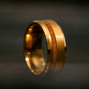 May include: A gold-colored ring with a brushed metal finish. The ring features a central band of light brown material, creating a two-tone effect. The ring is shown against a dark background.