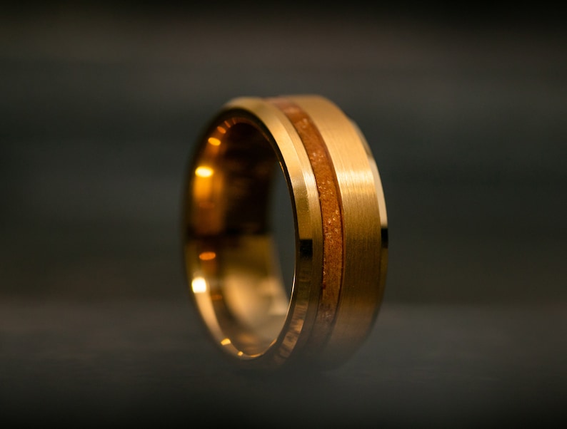 May include: A gold-colored ring with a brushed metal finish. The ring features a central band of light brown material, creating a two-tone effect. The ring is shown against a dark background.