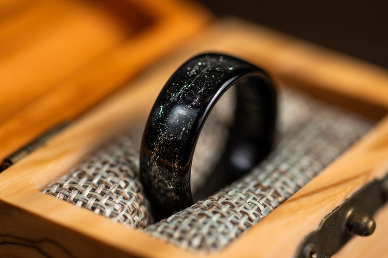 K&ouml;nnte beinhalten: Ein schwarzer Ring mit einem silbernen Streifen, der durch ihn verl&auml;uft, liegt in einer Holzschachtel mit Stoffauskleidung.