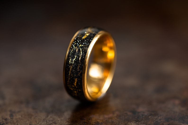 K&ouml;nnte beinhalten: Ein Goldring mit einer schwarzen Einlage mit goldenen Flocken. Der Ring hat eine polierte Goldoberfl&auml;che.