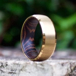 May include: A gold and wood ring with a dark wood inlay. The ring is on a gray surface.