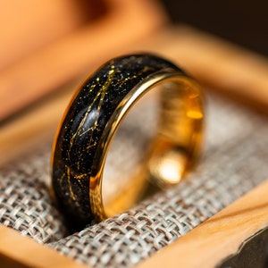 K&ouml;nnte beinhalten: Ein schwarzer und goldener Ehering mit einem goldenen Band und einem schwarzen Band mit goldenen Flocken. Der Ring befindet sich in einer Holzschachtel mit einem Stofffutter.