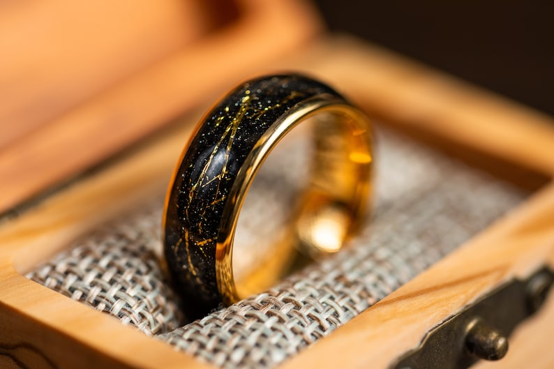 K&ouml;nnte beinhalten: Ein schwarzer und goldener Ehering mit einem goldenen Band und einem schwarzen Band mit goldenen Flocken. Der Ring befindet sich in einer Holzschachtel mit einem Stofffutter.