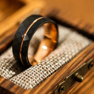 Puede incluir: Un anillo de carburo de tungsteno negro con una incrustaci&oacute;n de oro rosa y un acabado martillado. El anillo est&aacute; en una caja de madera con un forro de tela.