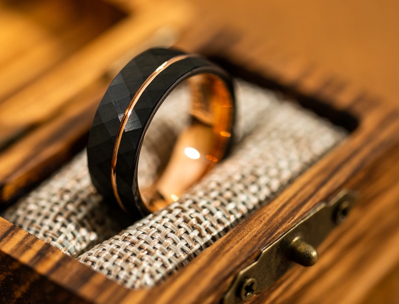 Puede incluir: Un anillo de carburo de tungsteno negro con una incrustaci&oacute;n de oro rosa y un acabado martillado. El anillo est&aacute; en una caja de madera con un forro de tela.