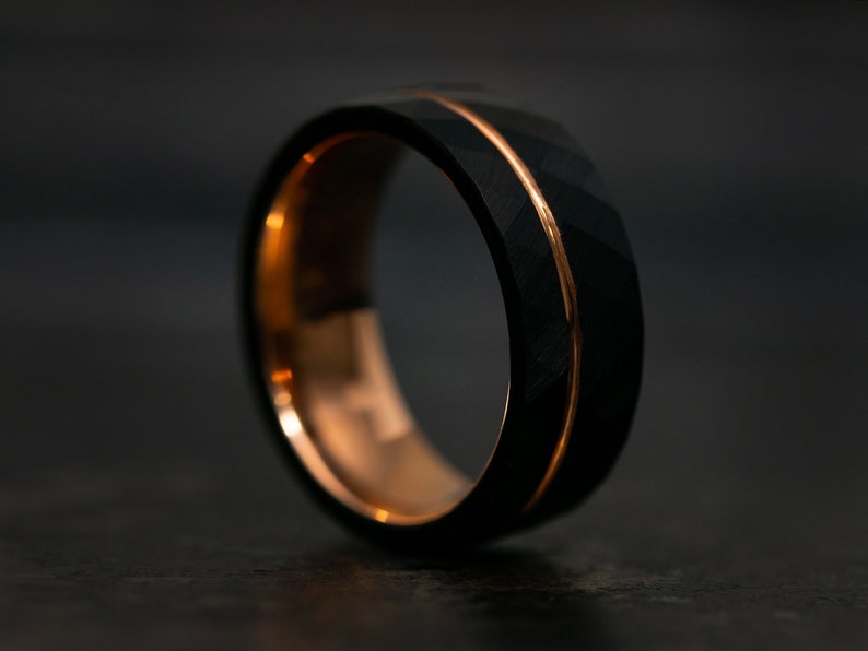 Puede incluir: Un anillo de carburo de tungsteno negro con una incrustaci&oacute;n de oro rosa. El anillo tiene un acabado martillado.