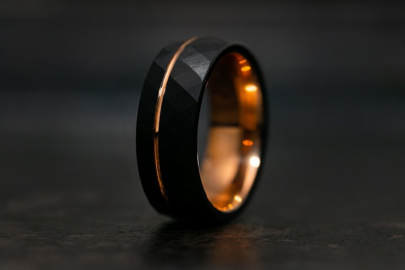 Puede incluir: Un anillo de carburo de tungsteno negro con una incrustaci&oacute;n de oro rosa. El anillo tiene un acabado cepillado y un borde biselado.