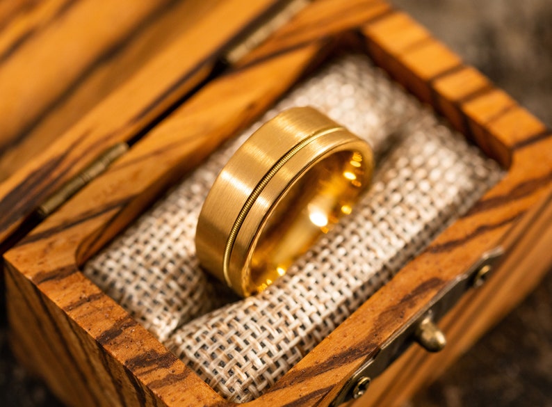 Puede incluir: Una banda de boda de oro con un acabado cepillado y una sola ranura alrededor del centro. El anillo est&aacute; en una caja de madera con un forro de arpillera.