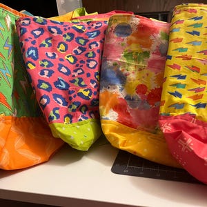 May include: A collection of colorful tote bags with various patterns. One bag features green fabric with yellow and orange lightning bolts, another has pink leopard print, and a third displays a watercolor design. The bags have contrasting colored bottoms.