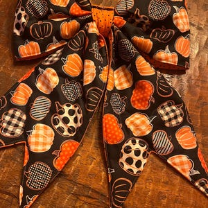 May include: A large fabric bow featuring a black background with a repeating pattern of orange pumpkins. The pumpkins display various designs including orange and white gingham, black and white polka dots, and solid orange. The center knot is orange.