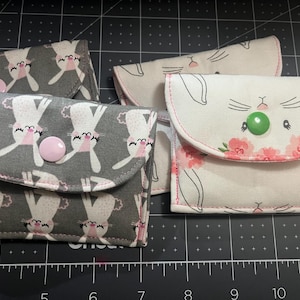 May include: Four fabric pouches with bunny face designs. Two pouches have a gray background with white bunnies and pink accents. The other two pouches have a white background with pink floral accents and a green button closure.