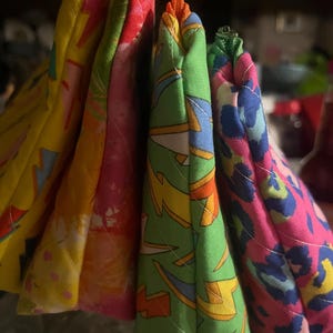 May include: A collection of colorful, quilted pot holders. The pot holders feature vibrant patterns including lightning bolts, floral designs, and animal prints. The pot holders are in shades of yellow, pink, green, and blue.