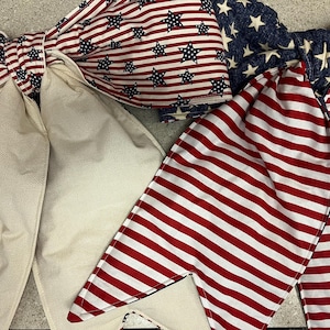 May include: Two decorative fabric bows featuring patriotic patterns. One bow displays red and white stripes with blue stars, paired with solid cream-colored tails. The second bow features a blue field with white stars and red and white striped tails.