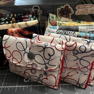 May include: A collection of fabric pouches with various patterns, including stethoscopes, floral designs, and equestrian themes. The pouches are white, black, and blue, with red accents and a snap closure. The pouches are stacked on a cutting mat.