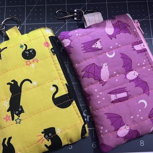 May include: Two rectangular zippered pouches with metal lobster claw clasps. The yellow pouch features a black cat pattern with small stars. The purple pouch features a bat and crescent moon pattern. Both pouches have horizontal quilted stitching.