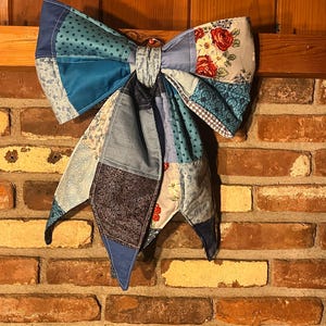 May include: A large decorative bow constructed from various quilted fabric patches in shades of blue, white, and red. The fabric features diverse patterns including small stars, floral rose prints, swirls, and geometric shapes, with two long hanging tails.