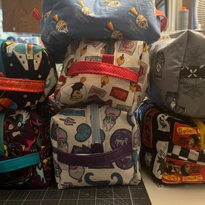 May include: A stack of small, square fabric pouches with zipper closures. The pouches feature various patterns, including cartoon villains, spiderwebs, and race cars. Each pouch has a different colored zipper and handle.