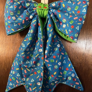 May include: A large fabric bow featuring a blue background with a repeating pattern of school supplies including globes, red apples, scissors, rulers, and glue bottles. The bow has a bright green fabric lining and two small red snaps at the center.