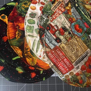 May include: A set of three circular fabric bowl covers featuring colorful harvest-themed prints. One cover has a black background with vegetables, another has a cream background with produce, and the third features vintage-style market labels.
