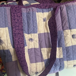 May include: A large, quilted tote bag in shades of purple, lavender, and white. The bag features a patchwork design with floral and polka dot patterns. The handles and trim are a darker purple with a floral print. The bag is designed for carrying items.