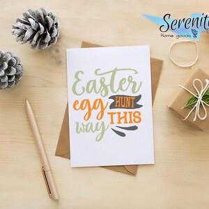 May include: A white card with the text "Easter egg hunt this way" in green, orange, and black lettering. The card is on a wooden surface with pine cones and a small wrapped gift.