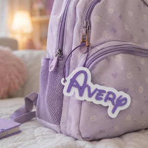 May include: A light purple backpack featuring a white and purple daisy and heart pattern. A white and purple custom name tag with the text AVERY is attached to the front zip pull with a thin cord. The backpack has a mesh side pocket.