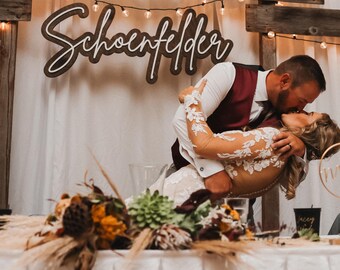 May include: A bride and groom kiss in front of a rustic wooden backdrop with a sign that reads "Schoenfelder". The sign is decorated with string lights. The couple is surrounded by a floral arrangement with dried grasses and succulents.