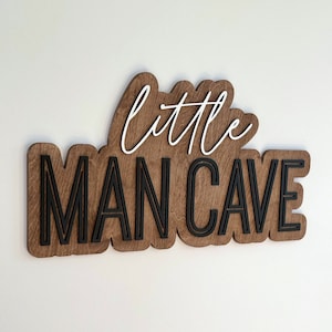 May include: A wooden sign with the words "little man cave" in black lettering on a brown background. The word "little" is written in white cursive.