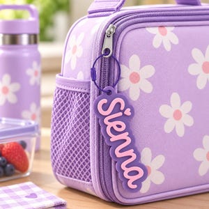 May include: A lavender lunch bag featuring a white daisy pattern with pink centres. A purple silicone luggage tag with the name Sienna in raised pink lettering is attached to the zip pull. A matching lavender water bottle is visible nearby.