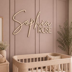 May include: Wooden name sign spelling "Sophia Rose" in a cursive font, mounted on a mauve wall. A light wood crib is in the foreground, with a teddy bear and pillow. The room is decorated in neutral tones.