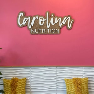 May include: A pink wall with a wooden sign that says "Carolina Nutrition" in white lettering. The sign is mounted on the wall above a white wave-patterned bench with two yellow pillows.