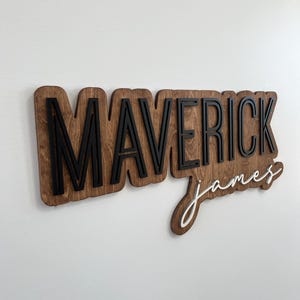 May include: A custom wooden wall sign featuring the name MAVERICK in bold, black, block capital letters layered over a dark stained wood backing, with the name james in white, cursive script lettering positioned below the main text.