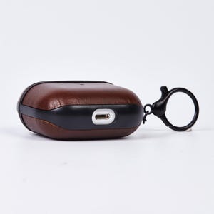 May include: A dark brown leather protective case for wireless earbuds featuring a black plastic frame and a black circular keychain attachment. The case includes a precise cutout on the bottom edge for a charging port, showcasing a smooth finish.