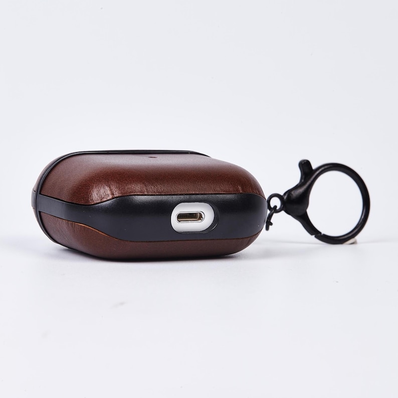 May include: A dark brown leather protective case for wireless earbuds featuring a black plastic frame and a black circular keychain attachment. The case includes a precise cutout on the bottom edge for a charging port, showcasing a smooth finish.