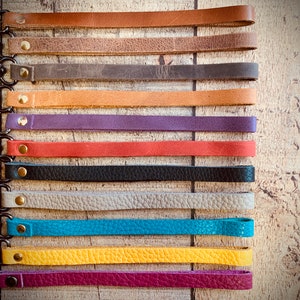 Op de afbeelding: Een verzameling leren polsbandjes in diverse kleuren, waaronder bruin, grijs, oranje, paars, rood, zwart, beige, turquoise, geel en magenta. Elke band heeft een metalen sluiting en een messing klinknagel.